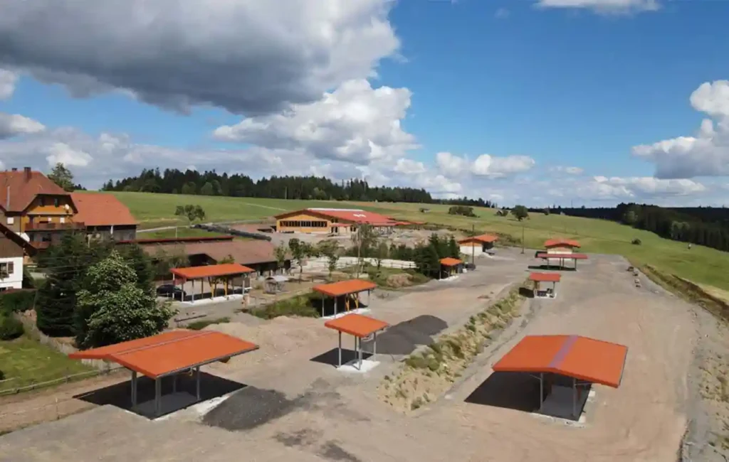Bilden visar en gård från fågelperspektiv. Flera små tak eller öppna skydd är utspridda över området. Till vänster i utkanten finns ett större hus. Hela området är omgivet av gröna ängar och träd och ligger på en lätt sluttning. Bilden ger en omfattande översikt över gården och dess utomhusområden. Principen för HIT-aktivstall: Rumslig åtskillnad mellan funktionsområdena marknadsplats (ligghall, foderautomater, fodertråg, vattenkälla) och strukturerade gångvägar eller stigar inkl. rullningsplats, dödträdshäck, hästtrappa, trädstamlabyrint, vattenfåra etc.