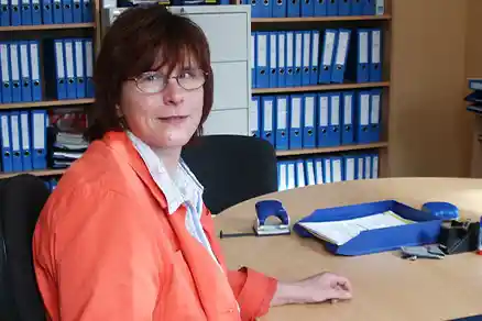 Kontaktpersoner 2 En kvinna sitter till vänster i bilden vid sin arbetsplats och tittar in i kameran. Bakom henne står flera hyllor som är fyllda med pärmar.